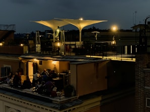 A rooftop terrace is dimly lit with warm, ambient lights during the evening. People are gathered around tables, engaged in conversation and enjoying a relaxed atmosphere. The terrace features modern outdoor furniture and large, white, triangular shade structures. The sky is dark, suggesting nighttime.
