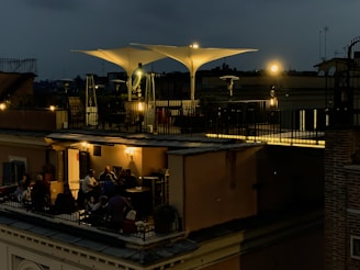 A rooftop terrace is dimly lit with warm, ambient lights during the evening. People are gathered around tables, engaged in conversation and enjoying a relaxed atmosphere. The terrace features modern outdoor furniture and large, white, triangular shade structures. The sky is dark, suggesting nighttime.