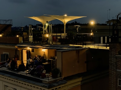 A rooftop terrace is dimly lit with warm, ambient lights during the evening. People are gathered around tables, engaged in conversation and enjoying a relaxed atmosphere. The terrace features modern outdoor furniture and large, white, triangular shade structures. The sky is dark, suggesting nighttime.