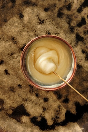 A ceramic bowl containing a thick, creamy substance, possibly honey or some type of paste. The substance has a smooth texture and is being stirred with a thin wooden stick. The bowl rests on a textured, speckled surface in shades of beige and brown.