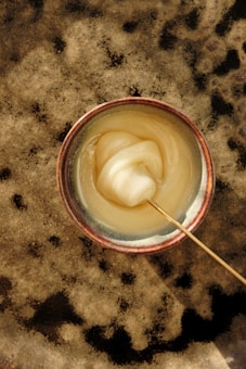 A ceramic bowl containing a thick, creamy substance, possibly honey or some type of paste. The substance has a smooth texture and is being stirred with a thin wooden stick. The bowl rests on a textured, speckled surface in shades of beige and brown.