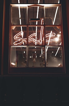 A window with a neon sign displaying the word 'sale' in cursive script. The neon light is bright, with reflections visible on the window panes. The interior lighting creates a warm and inviting ambiance, with some industrial-style bulbs hanging in the background.