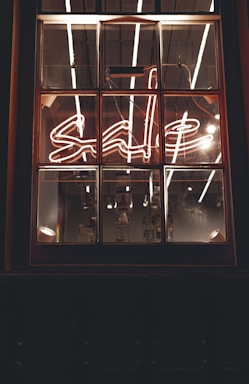 A window with a neon sign displaying the word 'sale' in cursive script. The neon light is bright, with reflections visible on the window panes. The interior lighting creates a warm and inviting ambiance, with some industrial-style bulbs hanging in the background.
