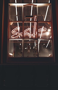 A window with a neon sign displaying the word 'sale' in cursive script. The neon light is bright, with reflections visible on the window panes. The interior lighting creates a warm and inviting ambiance, with some industrial-style bulbs hanging in the background.