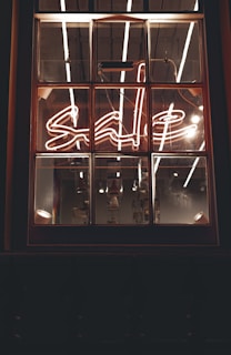 A window with a neon sign displaying the word 'sale' in cursive script. The neon light is bright, with reflections visible on the window panes. The interior lighting creates a warm and inviting ambiance, with some industrial-style bulbs hanging in the background.