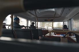 The interior of a public bus with several passengers seated, viewed from the back towards the front. The scene appears to be somewhat dimly lit, with light coming in from the large windows. Signs and a digital display show bus route information. The passengers are dressed warmly, suggesting cooler weather outside.