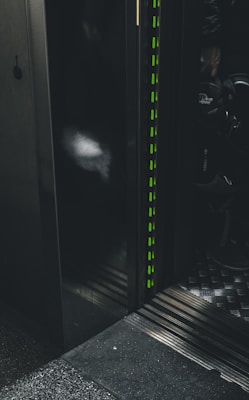 A dimly lit elevator interior with metal paneling and a striped pattern on the floor. The edge of the elevator door features vertical green lights, and some shadows are visible on the wall.