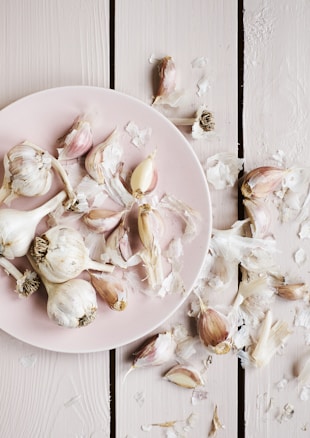 Several bulbs and cloves of garlic are arranged on a light pink plate set on a wooden surface. The scene includes scattered garlic skins around the plate and on the table, creating a rustic and natural aesthetic.