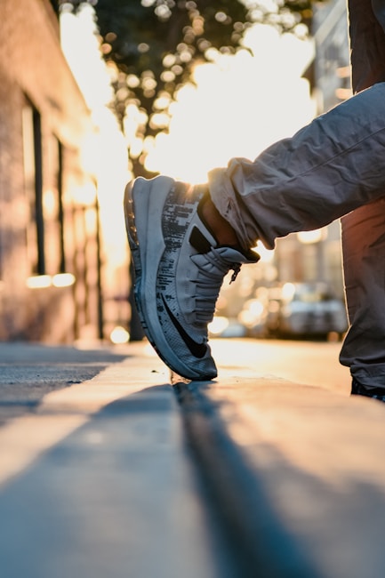 A close-up of a person wearing sneakers with one foot resting on the edge of a sidewalk. The scene is set during sunset or sunrise, with a warm glow in the background and soft lighting. The street and buildings are slightly blurred, enhancing the focus on the shoes.