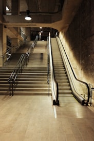 An interior space featuring a set of stairs and an escalator side by side, leading upwards. The area is illuminated by overhead industrial-style lights and has concrete walls and floors, creating a utilitarian and modern vibe.