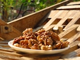 A white plate filled with walnuts is placed on a wooden surface with an intricate pattern. The background features some greenery, suggesting an outdoor setting. The scene is bathed in warm, golden sunlight, highlighting the texture of the walnuts and wood.