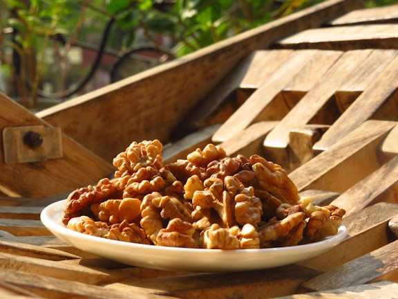 A white plate filled with walnuts is placed on a wooden surface with an intricate pattern. The background features some greenery, suggesting an outdoor setting. The scene is bathed in warm, golden sunlight, highlighting the texture of the walnuts and wood.