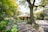 A serene garden leads up to a quaint house surrounded by lush greenery and vibrant flowers. A large tree dominates the foreground, with people walking along a path in the background. The scene conveys tranquility and natural beauty.