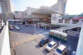 A bustling train station area with several parked cars and taxis lining the street. A green and beige bus is passing by near the entrance. The station building features a large entrance with an overhang and a prominent station sign. Pedestrian walkways and shops are visible along the station's facade.