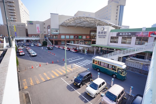 A bustling train station area with several parked cars and taxis lining the street. A green and beige bus is passing by near the entrance. The station building features a large entrance with an overhang and a prominent station sign. Pedestrian walkways and shops are visible along the station's facade.