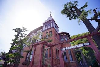 A historic-looking, red-brick building with intricate architectural details, including tall windows and a pointed roof. A wrought iron archway with the word 'Rhenania' is prominently displayed in front of the building. Surrounding the building are tall leafy trees, suggesting a serene and stately atmosphere.