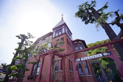 A historic-looking, red-brick building with intricate architectural details, including tall windows and a pointed roof. A wrought iron archway with the word 'Rhenania' is prominently displayed in front of the building. Surrounding the building are tall leafy trees, suggesting a serene and stately atmosphere.