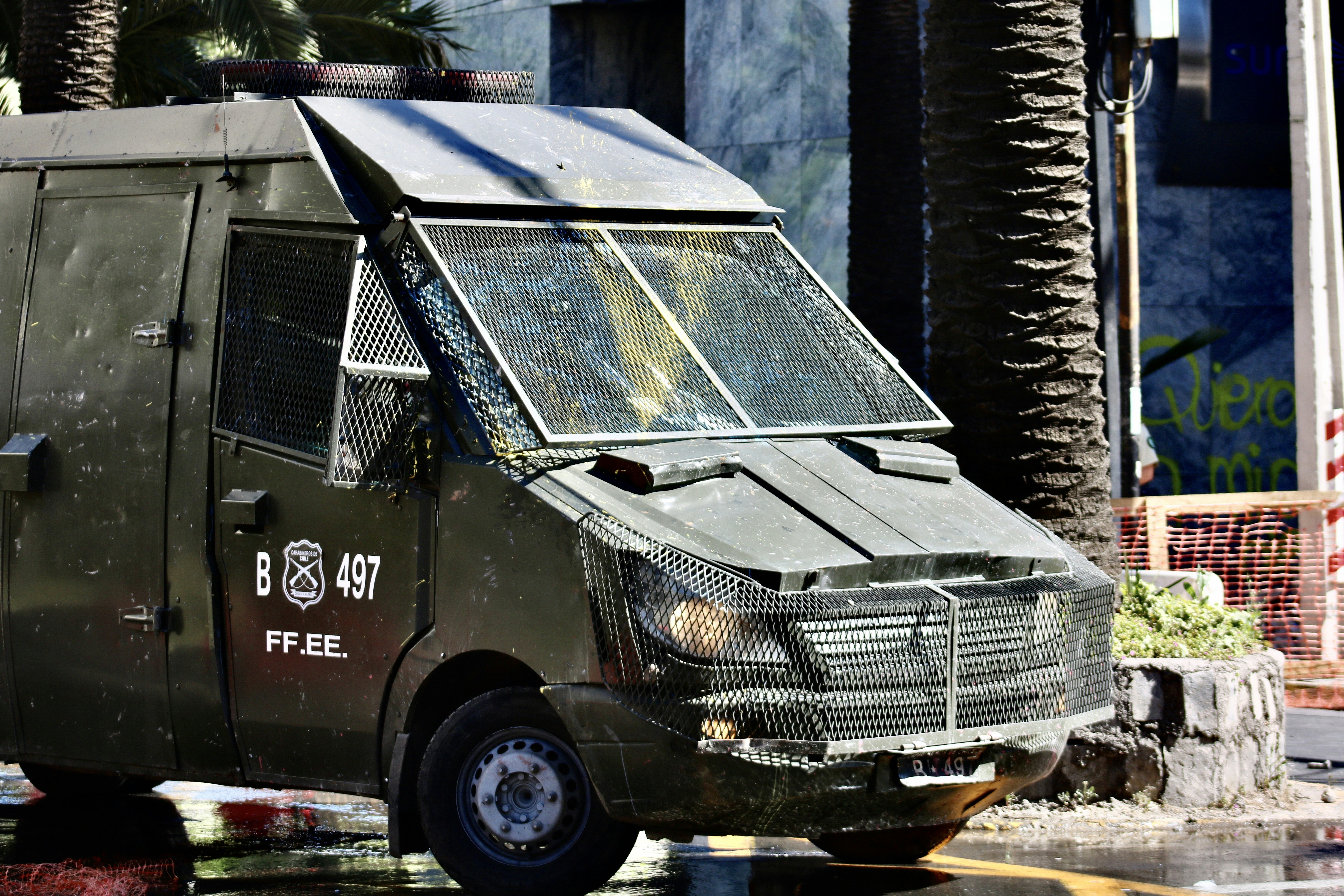 A heavily armored police vehicle is parked on a city street near palm trees and a stone planter. The vehicle has protective metal grating over the windows and visible markings with the letters 'B 497' and 'FF.EE.'. The surrounding environment includes urban elements such as a sidewalk, a stone planter, and construction fencing.