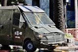 A heavily armored police vehicle is parked on a city street near palm trees and a stone planter. The vehicle has protective metal grating over the windows and visible markings with the letters 'B 497' and 'FF.EE.'. The surrounding environment includes urban elements such as a sidewalk, a stone planter, and construction fencing.