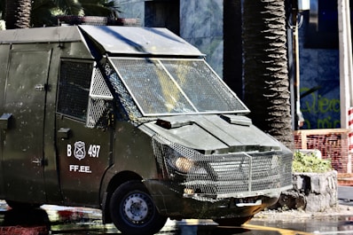 A heavily armored police vehicle is parked on a city street near palm trees and a stone planter. The vehicle has protective metal grating over the windows and visible markings with the letters 'B 497' and 'FF.EE.'. The surrounding environment includes urban elements such as a sidewalk, a stone planter, and construction fencing.