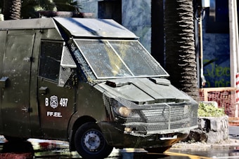 A heavily armored police vehicle is parked on a city street near palm trees and a stone planter. The vehicle has protective metal grating over the windows and visible markings with the letters 'B 497' and 'FF.EE.'. The surrounding environment includes urban elements such as a sidewalk, a stone planter, and construction fencing.