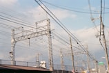 Metallic electrical towers and poles supporting numerous power lines stretch across a cloudy sky. The structures are intricate and industrial, emphasizing the complex network of cables overhead. The scenery includes elements of urban infrastructure like rooftops and a few signs.
