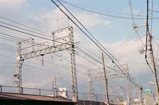 Metallic electrical towers and poles supporting numerous power lines stretch across a cloudy sky. The structures are intricate and industrial, emphasizing the complex network of cables overhead. The scenery includes elements of urban infrastructure like rooftops and a few signs.