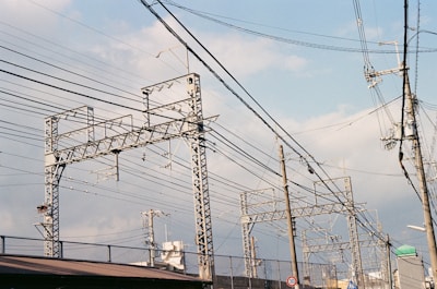 Metallic electrical towers and poles supporting numerous power lines stretch across a cloudy sky. The structures are intricate and industrial, emphasizing the complex network of cables overhead. The scenery includes elements of urban infrastructure like rooftops and a few signs.