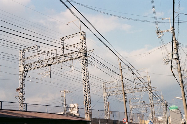 Metallic electrical towers and poles supporting numerous power lines stretch across a cloudy sky. The structures are intricate and industrial, emphasizing the complex network of cables overhead. The scenery includes elements of urban infrastructure like rooftops and a few signs.