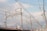 Metallic electrical towers and poles supporting numerous power lines stretch across a cloudy sky. The structures are intricate and industrial, emphasizing the complex network of cables overhead. The scenery includes elements of urban infrastructure like rooftops and a few signs.