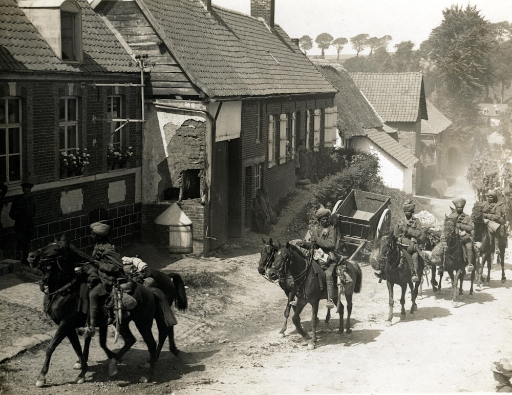 gente montando a caballo al lado de las casas fotografía en escala de grises