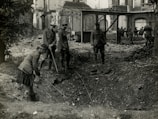 Four soldiers examine a large crater in a war-torn area, surrounded by debris and damaged buildings. They are dressed in military uniforms and appear to be using sticks or poles to inspect the crater. The scene is chaotic, indicating the aftermath of a violent event.