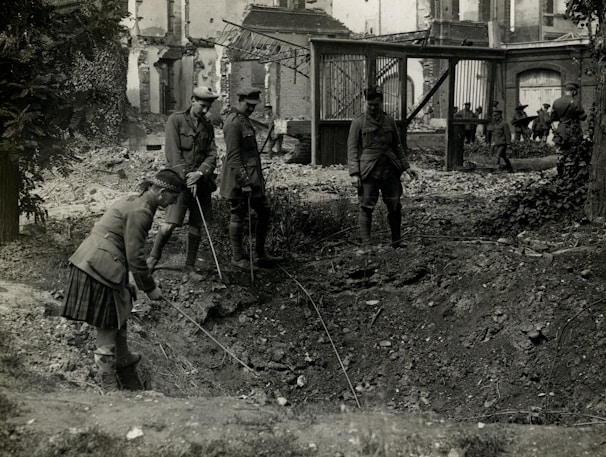 Four soldiers examine a large crater in a war-torn area, surrounded by debris and damaged buildings. They are dressed in military uniforms and appear to be using sticks or poles to inspect the crater. The scene is chaotic, indicating the aftermath of a violent event.