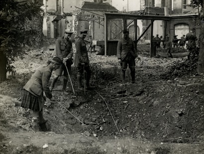 Four soldiers examine a large crater in a war-torn area, surrounded by debris and damaged buildings. They are dressed in military uniforms and appear to be using sticks or poles to inspect the crater. The scene is chaotic, indicating the aftermath of a violent event.