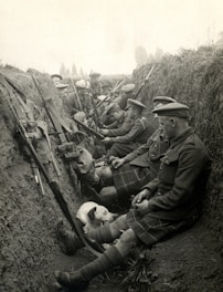 A group of soldiers in a trench, dressed in military uniforms and kilts, are seated and equipped with rifles. They appear to be resting, with some interacting with each other. A dog is also present, lying beside the soldiers.