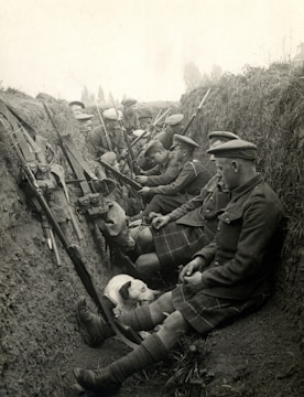 A group of soldiers in a trench, dressed in military uniforms and kilts, are seated and equipped with rifles. They appear to be resting, with some interacting with each other. A dog is also present, lying beside the soldiers.