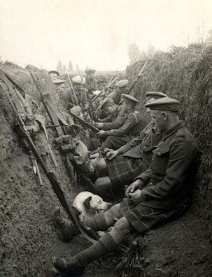 A group of soldiers in a trench, dressed in military uniforms and kilts, are seated and equipped with rifles. They appear to be resting, with some interacting with each other. A dog is also present, lying beside the soldiers.
