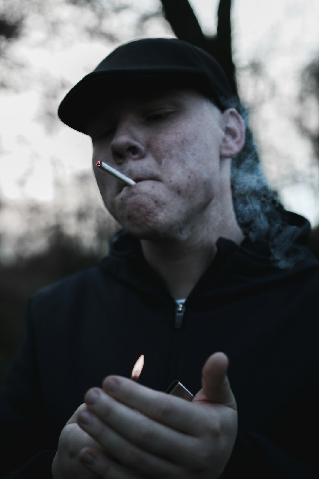 man wearing black zip-up jacket and black hat smoking cigarette stick while holding lighter