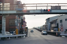 An urban street scene features an elevated pedestrian bridge with traffic signals attached. Various road signs are visible along the road, which is leading up a slight incline. On the left side, several buildings and parked vehicles are present. A few cars are driving up and down the road.
