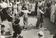 A group of couples are dancing closely in an outdoor setting, surrounded by a large crowd of onlookers dressed in mid-20th-century attire. The dancers appear to be engaged in a social or community event.