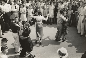 A group of couples are dancing closely in an outdoor setting, surrounded by a large crowd of onlookers dressed in mid-20th-century attire. The dancers appear to be engaged in a social or community event.
