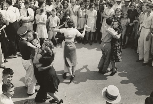 A group of couples are dancing closely in an outdoor setting, surrounded by a large crowd of onlookers dressed in mid-20th-century attire. The dancers appear to be engaged in a social or community event.