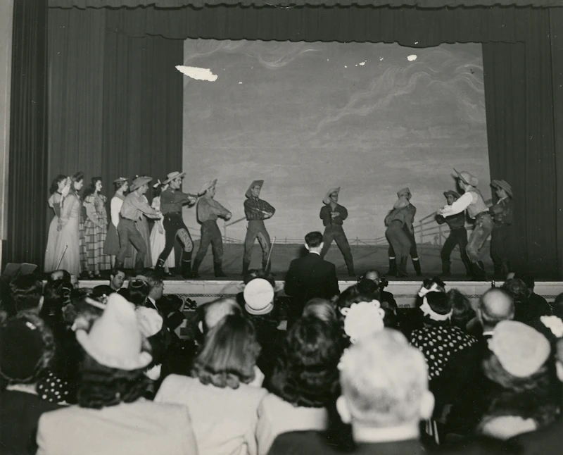 Archival photograph of a stage performance watched by a seated audience in hats