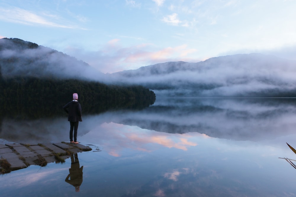 person standing on dock viewing mountain and body of water under blue and white sky