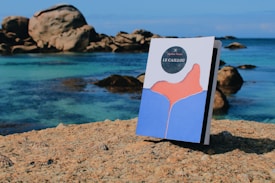 A book titled 'Le Caillou' stands upright on a rocky surface by the sea. The artwork on the cover features an abstract design with blue and pink shapes. In the background, large rocks and clear turquoise water are visible, creating a serene coastal scene under a clear blue sky.