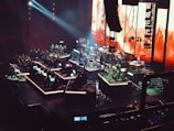 An orchestral performance taking place on a large stage with multiple musicians. The stage is illuminated with vibrant lights, featuring large screens displaying abstract images. The musicians are seated in sections with various instruments, including strings, brass, and percussion.