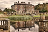 A large, old stone mansion with multiple chimneys and ivy-covered walls is situated in a landscaped garden. A tranquil pond in the foreground reflects the building's architecture. Stone balustrades border the water, and lush greenery surrounds the estate, with trees and manicured lawns adding to the scenic beauty.