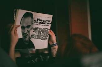A person is holding a magazine featuring a black-and-white photograph and text about a man. The layout includes large text in a bold font and a stylized portrait of the individual. The person's hands are visible, with a tattoo on the wrist and a wristband.