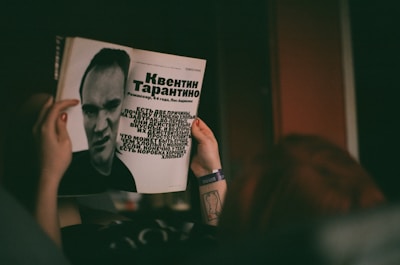 A person is holding a magazine featuring a black-and-white photograph and text about a man. The layout includes large text in a bold font and a stylized portrait of the individual. The person's hands are visible, with a tattoo on the wrist and a wristband.