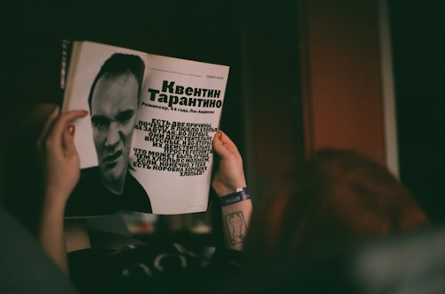 A person is holding a magazine featuring a black-and-white photograph and text about a man. The layout includes large text in a bold font and a stylized portrait of the individual. The person's hands are visible, with a tattoo on the wrist and a wristband.